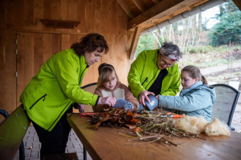 Twee volwassenen en twee kinderen maken iets met bladeren en takken aan een tafel buiten.