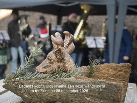 Hertenbeeld in kerstversiering met muzikanten op achtergrond, tekst over wandeltocht op 26 december 2026.