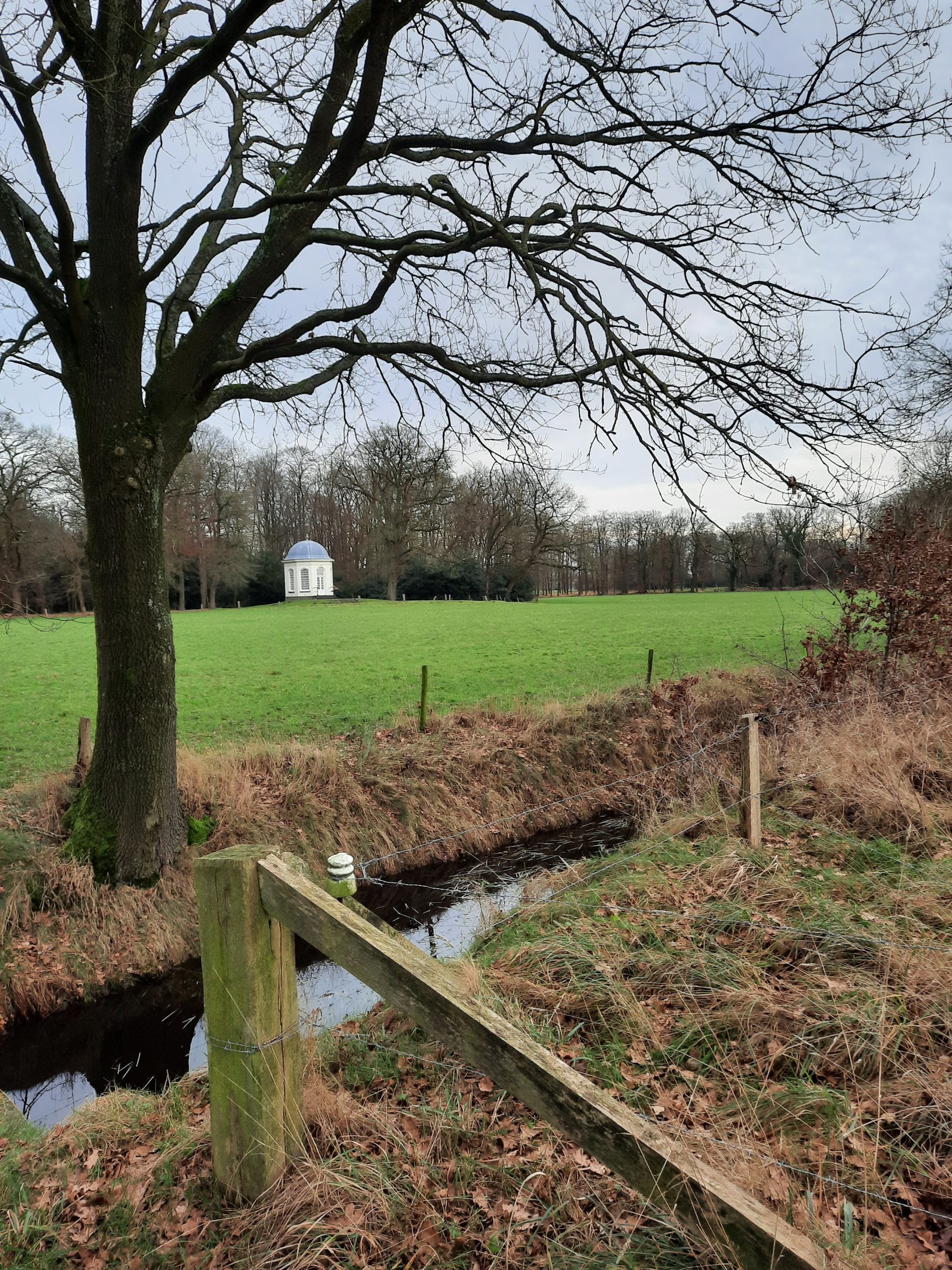 Groene weide met beek, bomen en een wit prieel op de achtergrond onder een bewolkte hemel.