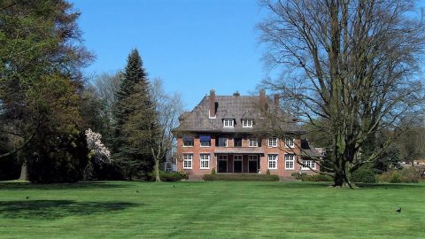 Groot bakstenen huis met een grasveld en bomen, onder een heldere blauwe lucht.