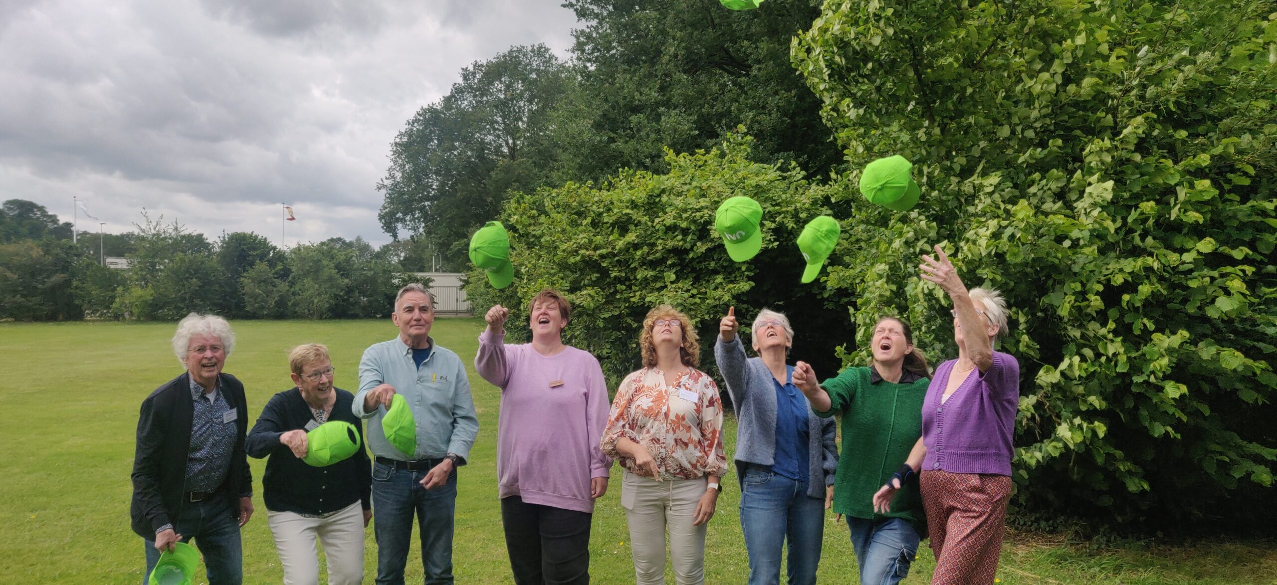 Een groep mensen gooit groene petten in de lucht op een grasveld bij bomen.
