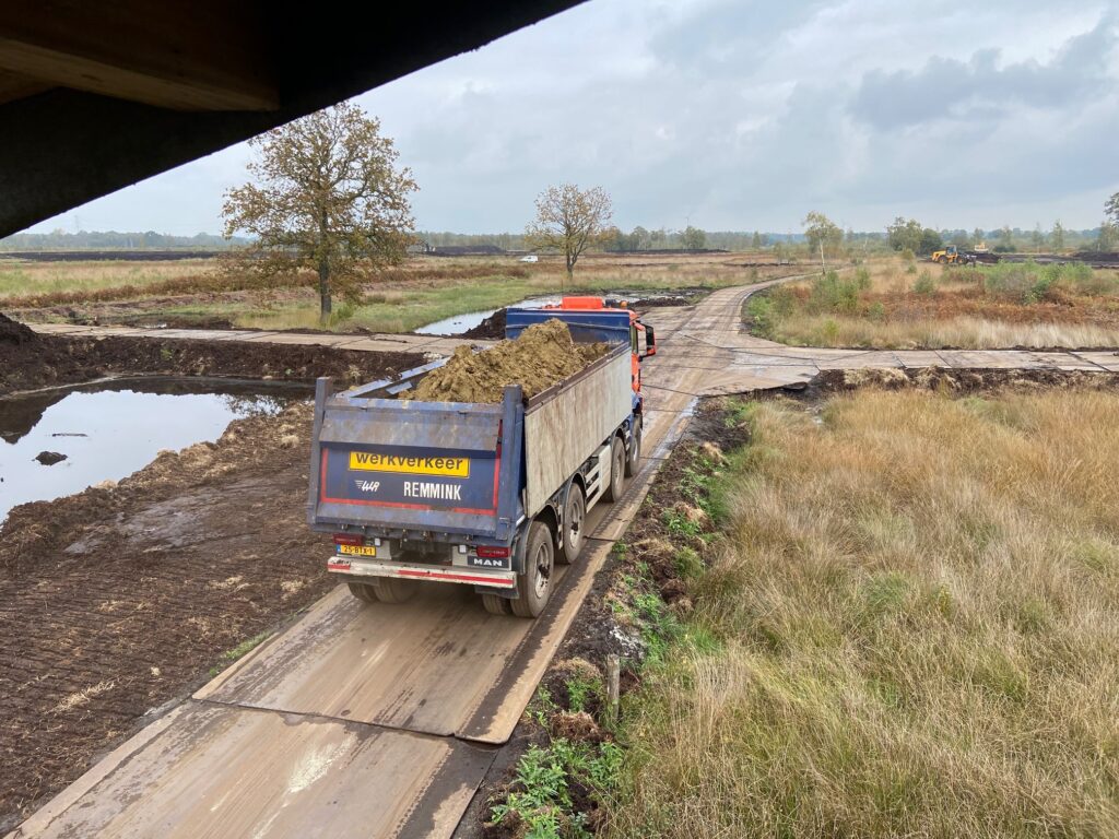 Vrachtwagen vervoert zand over modderige weg in landelijk gebied met grote plassen en groen.