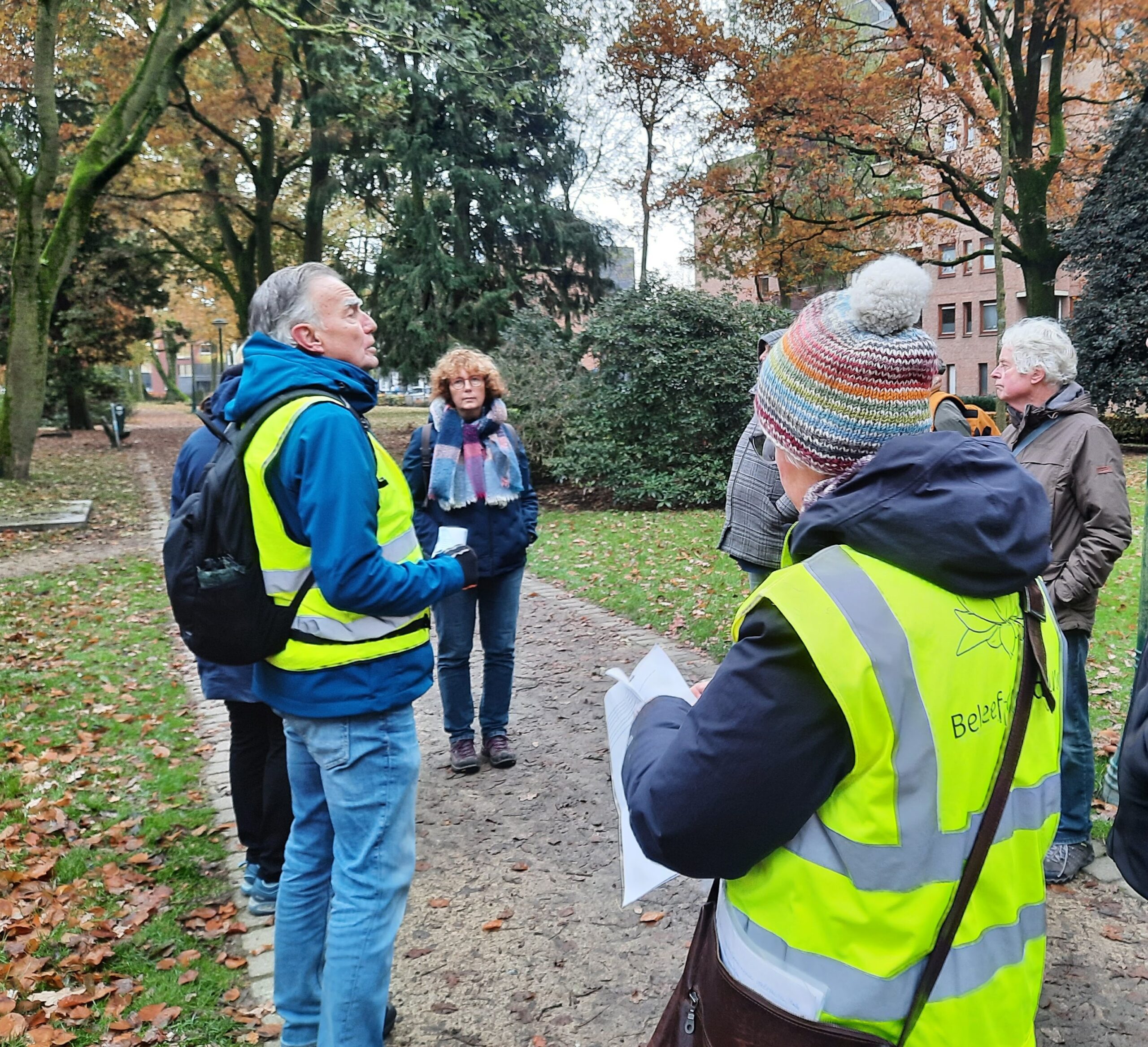 Groep mensen met gele hesjes in een herfstig park, in gesprek op een met bladeren bedekte pad.