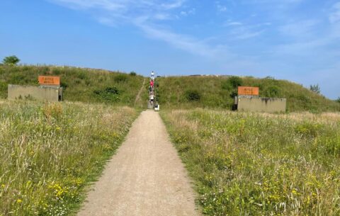Een pad leidt naar een heuvel met opstap en mensen, omringd door gras en twee structuren.