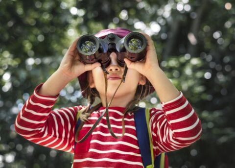 Kind in rood-wit gestreepte trui kijkt door een verrekijker in een groene omgeving.