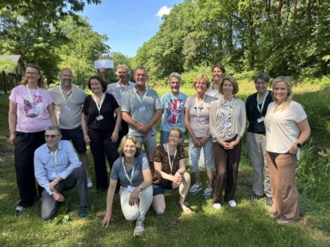 Een groep mensen poseert buiten in een groene, bosrijke omgeving op een zonnige dag.