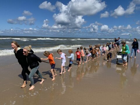 Groep kinderen en volwassenen maakt een menselijke ketting op een zonnig strand met zee op de achtergrond.