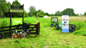 Ingang van Heemtuin De Dwarsgouw, met fietsenrekken en informatieborden, omgeven door gras en bomen.