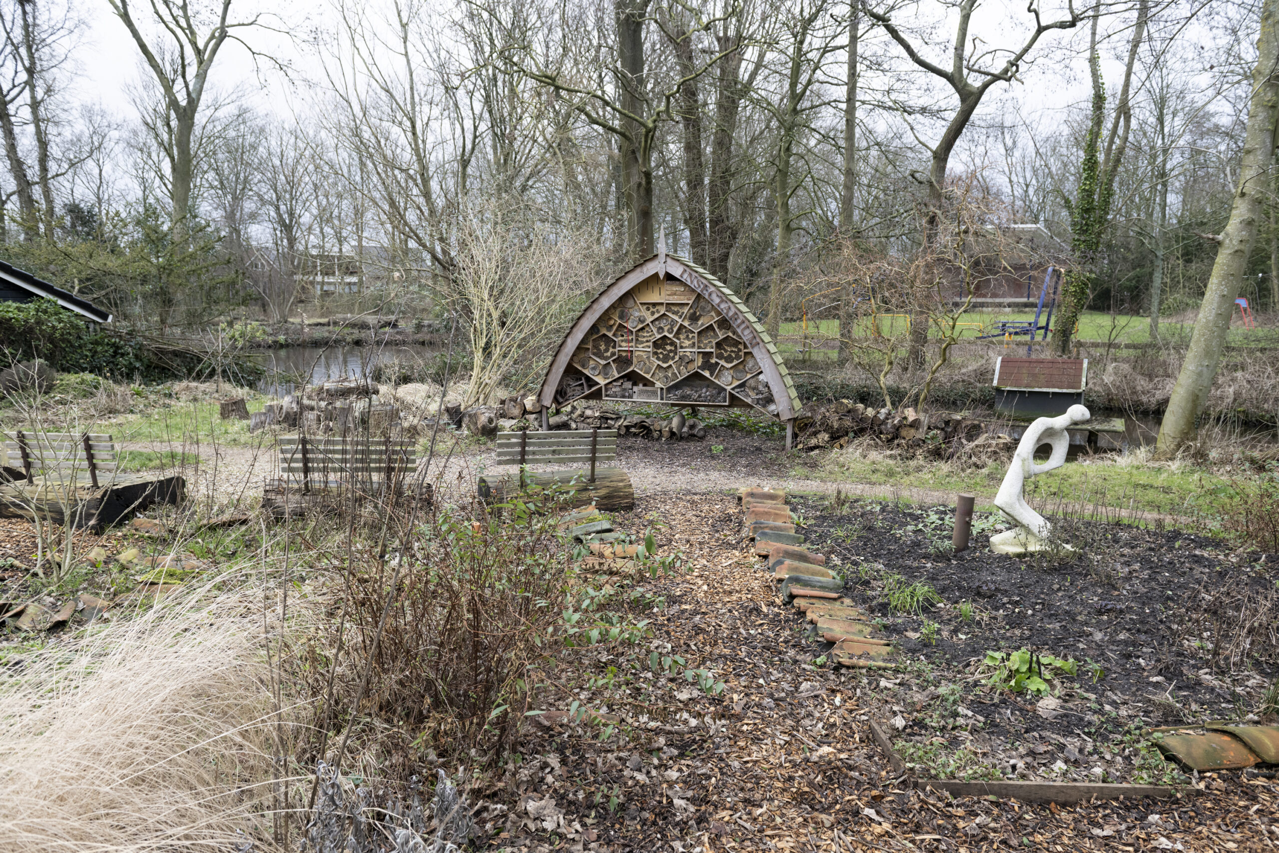 Tuin met insectenhotel, bankjes en wit beeld, omringd door bomen en struiken in een parkachtige omgeving.
