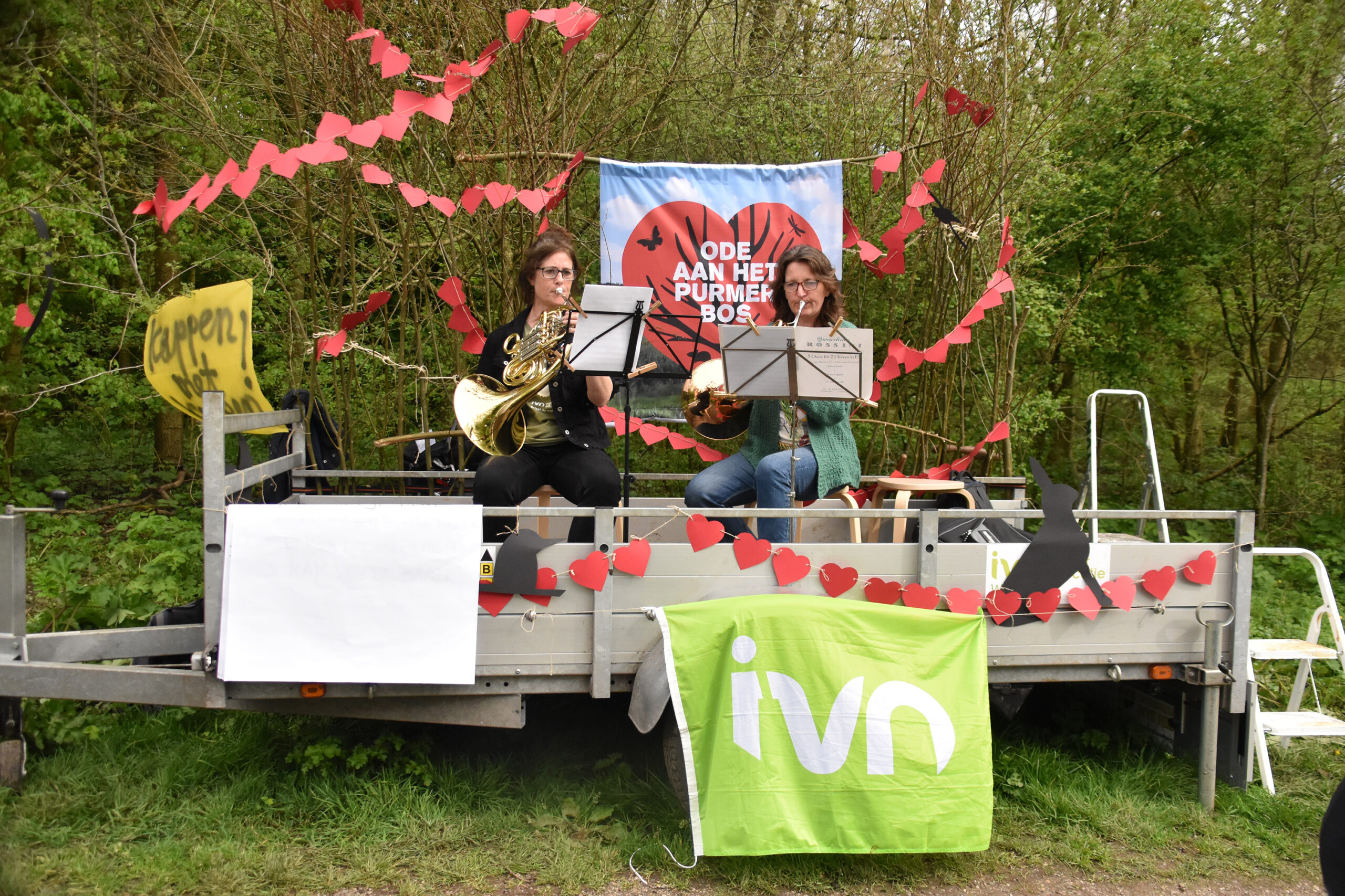 Twee muzikanten spelen hoorn op een versierde aanhanger in een bosrijke omgeving.