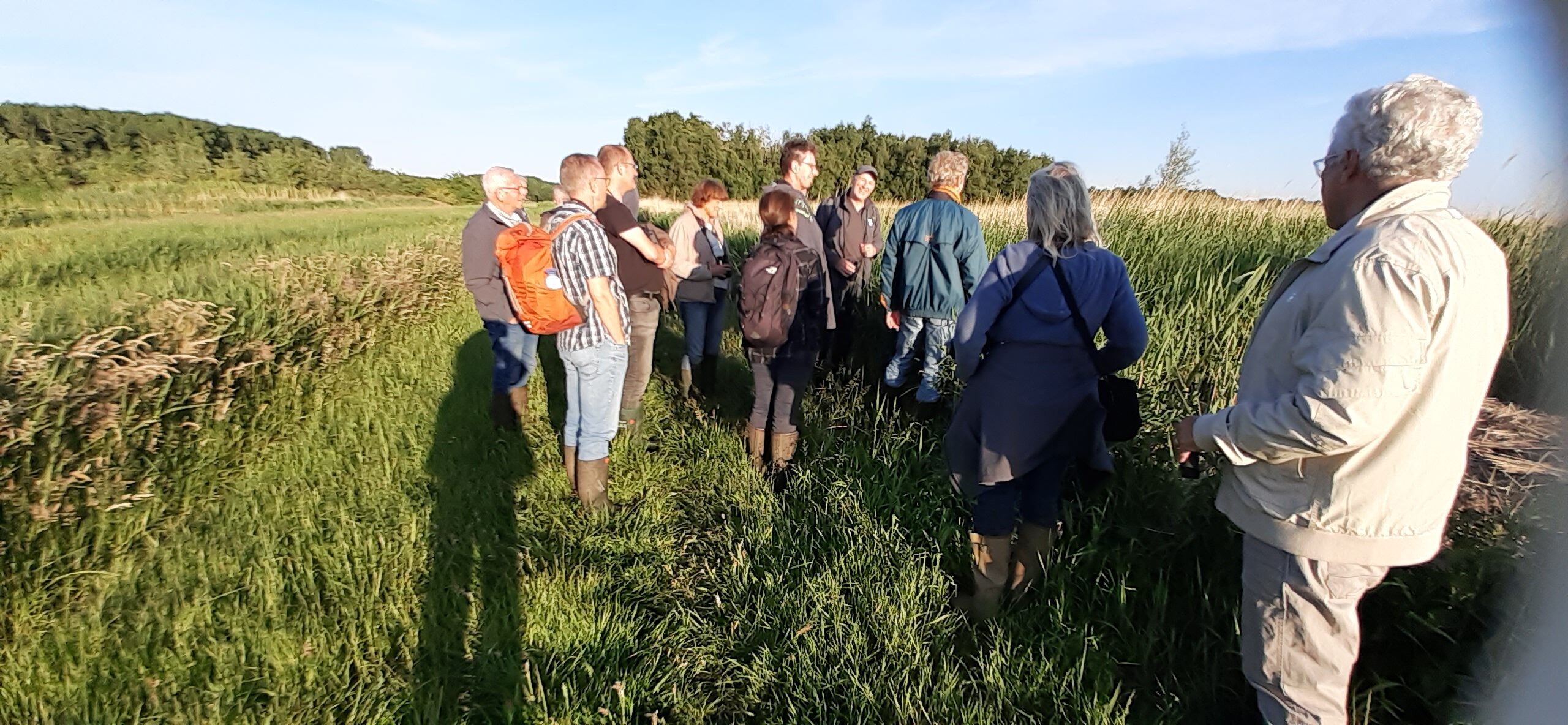 Groep mensen met rugzakken op een wandelpad in een groen veld met bomen en blauwe lucht.