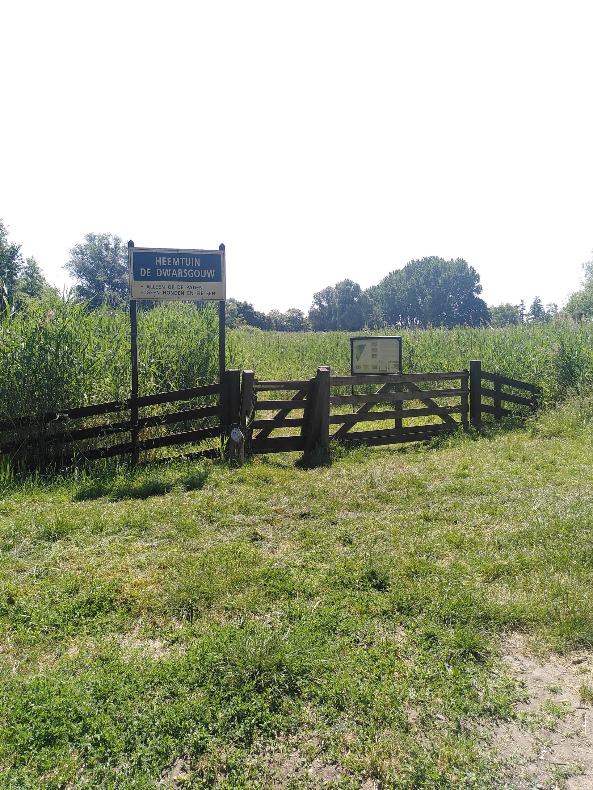 Toegangshek van Heemtuin De Dwarsgouw met bord. Weelderig groen en bomen op de achtergrond.