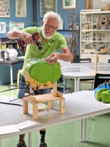 Man in groen shirt boort in een groen model in een klaslokaal met boeken en natuurposters.