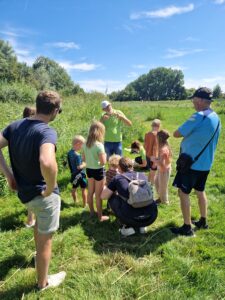 Groep mensen in een veld, aandachtig luisterend naar een spreker onder blauwe lucht.