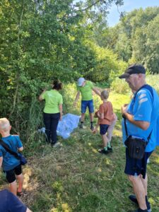 Mensen in groene shirts onderzoeken planten bij bosrand, begeleid door kinderen op zonnige dag.