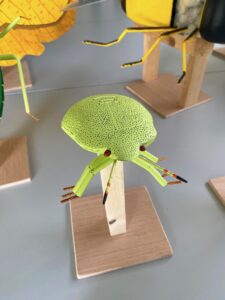 Groene insectensculptuur met stippen, staande op een houten standaard op een grijze tafel.