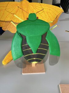 Groot model van een groene kever met gele vleugels, gepresenteerd op een houten standaard.