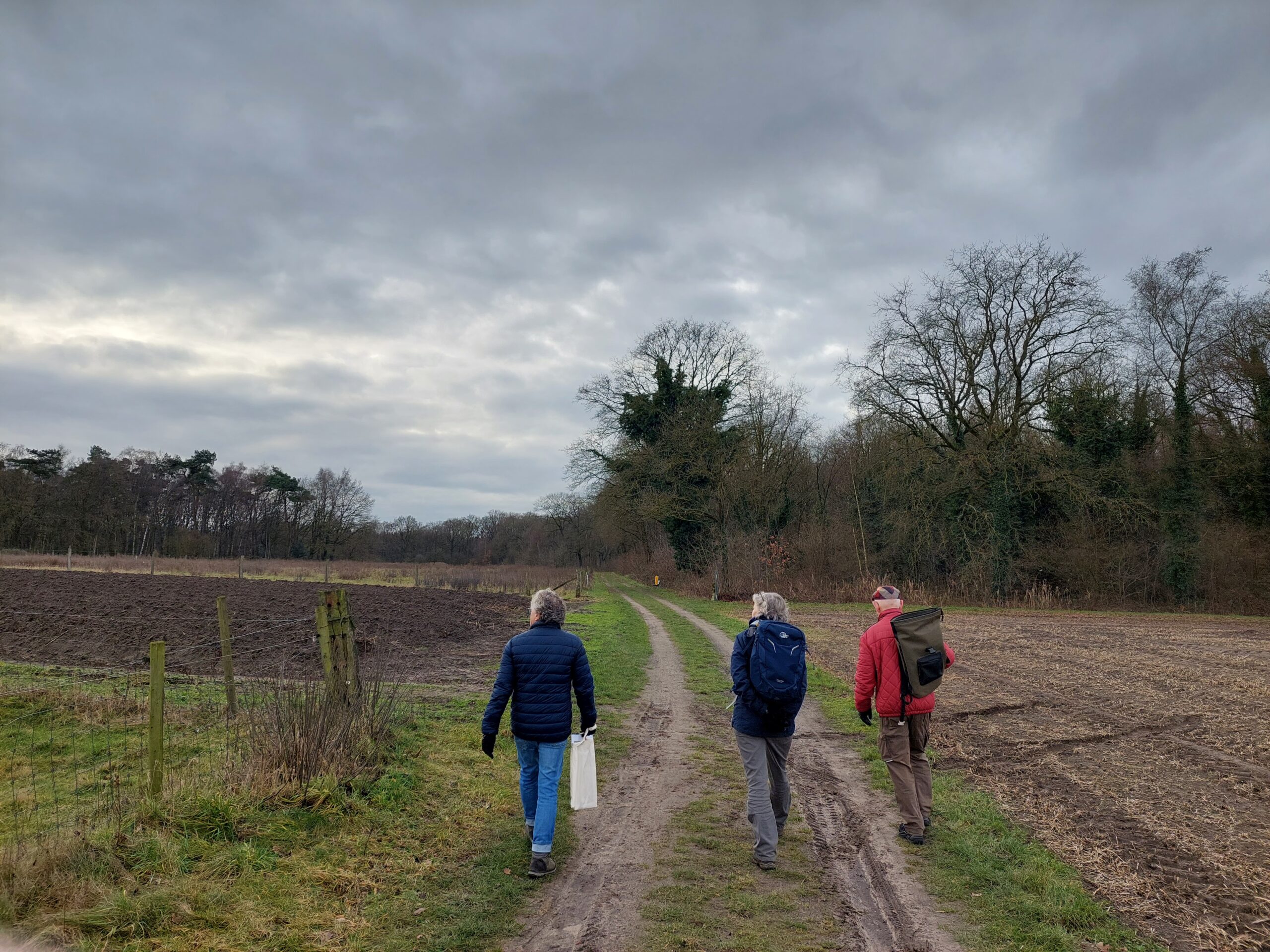 Drie mensen wandelen op een modderig pad langs velden onder een bewolkte hemel.