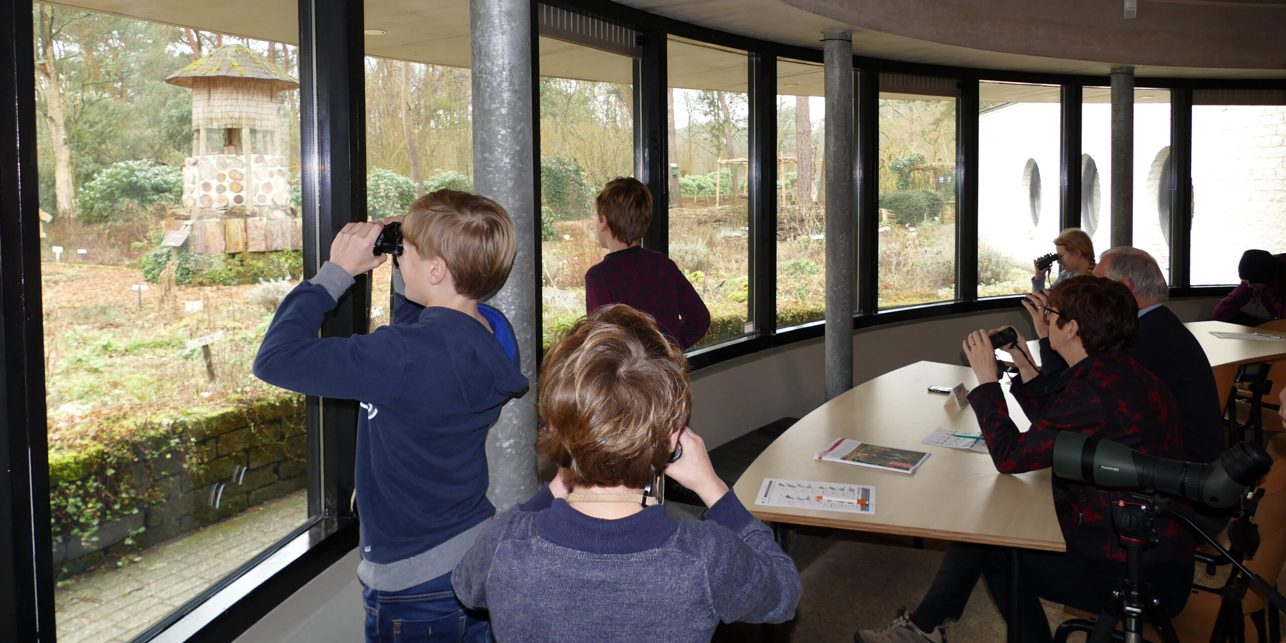 Mensen kijken met verrekijkers uit een observatieruimte naar een natuurlijke tuin.