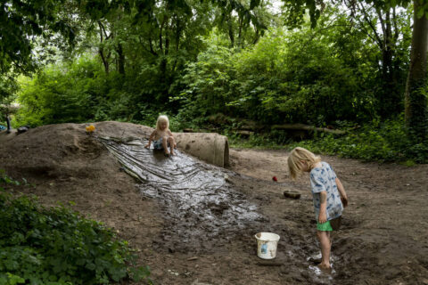 Twee kinderen spelen in de modder bij een plastic glijbaan in een bosrijke omgeving.