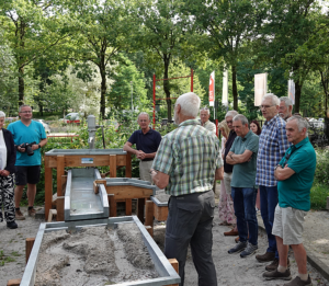 Een groep mensen luistert naar een uitleg bij een demonstratiemodel in een groene buitenomgeving.