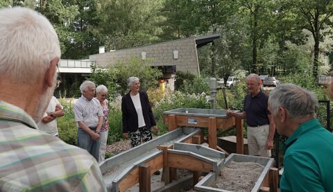 Groep oudere mensen buiten bij een zand- en waterbak, omgeven door groen en een modern gebouw.