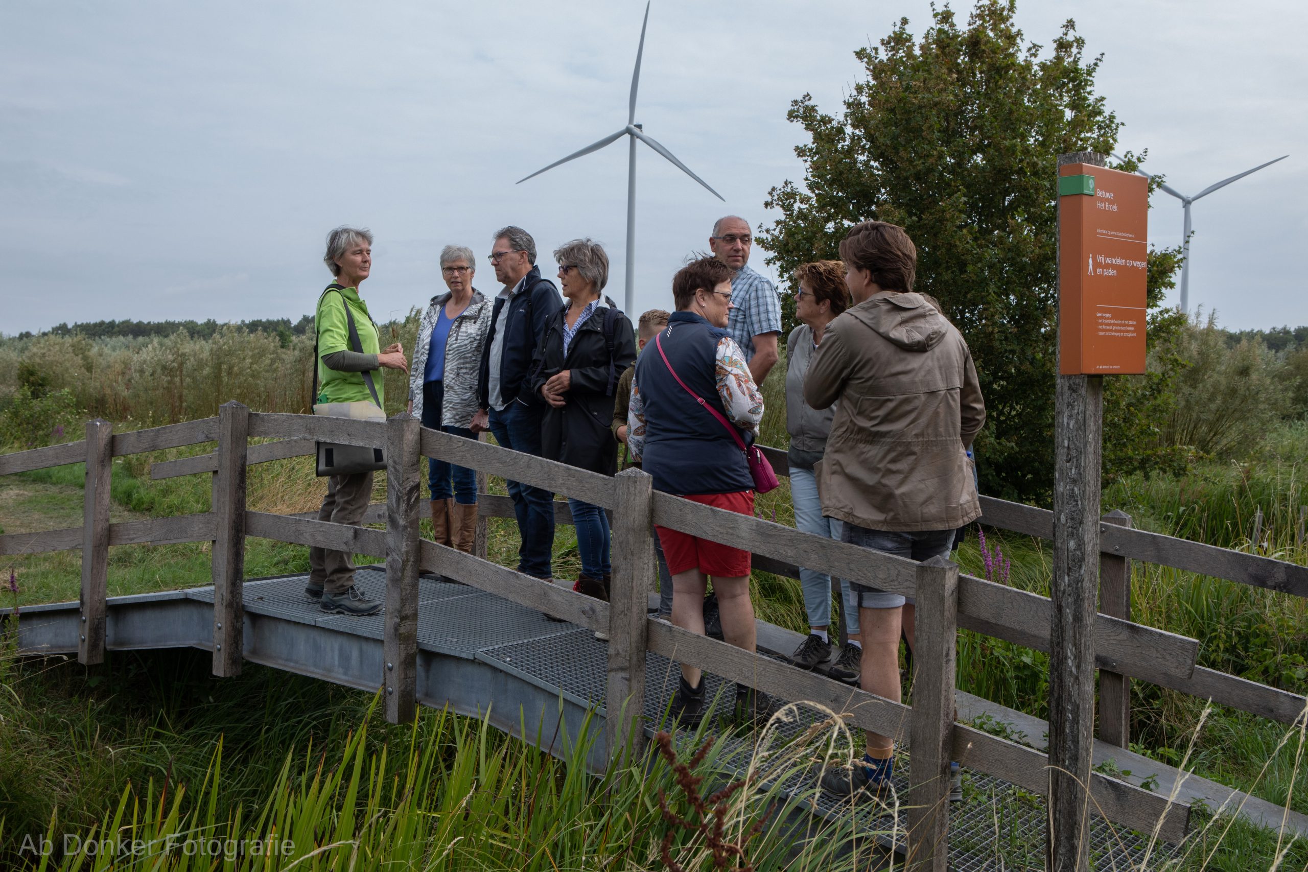 Groep mensen staat op een houten brug in een natuurgebied met een windmolen en een informatiebord.