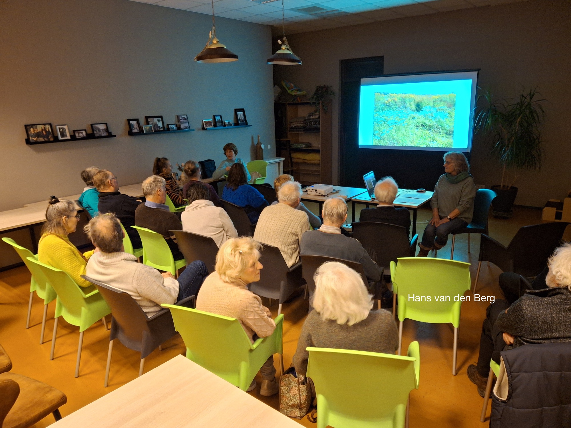 Een groep mensen luistert naar een presentatie met een natuurfoto op een projectiescherm in een zaal.