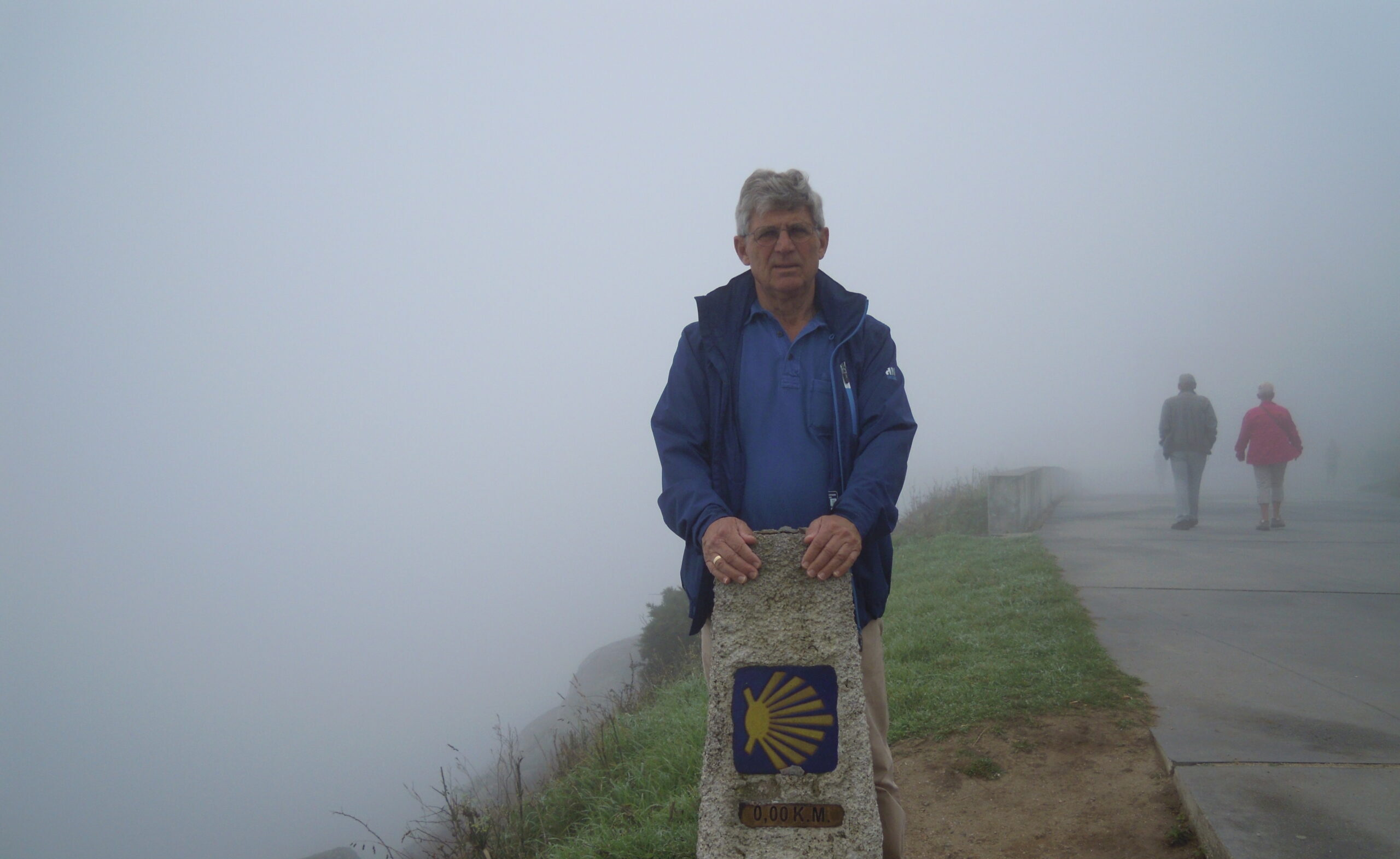 Man bij 0,00 km marker Camino de Santiago in mist, met wandelpad en mensen op achtergrond.
