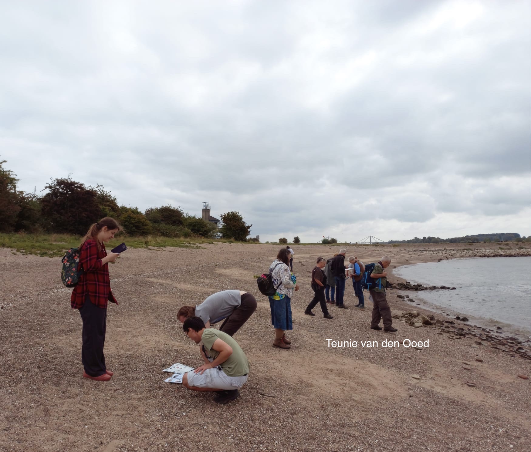 Een groep mensen onderzoekt een kiezelstrand langs een rivier onder een bewolkte hemel.