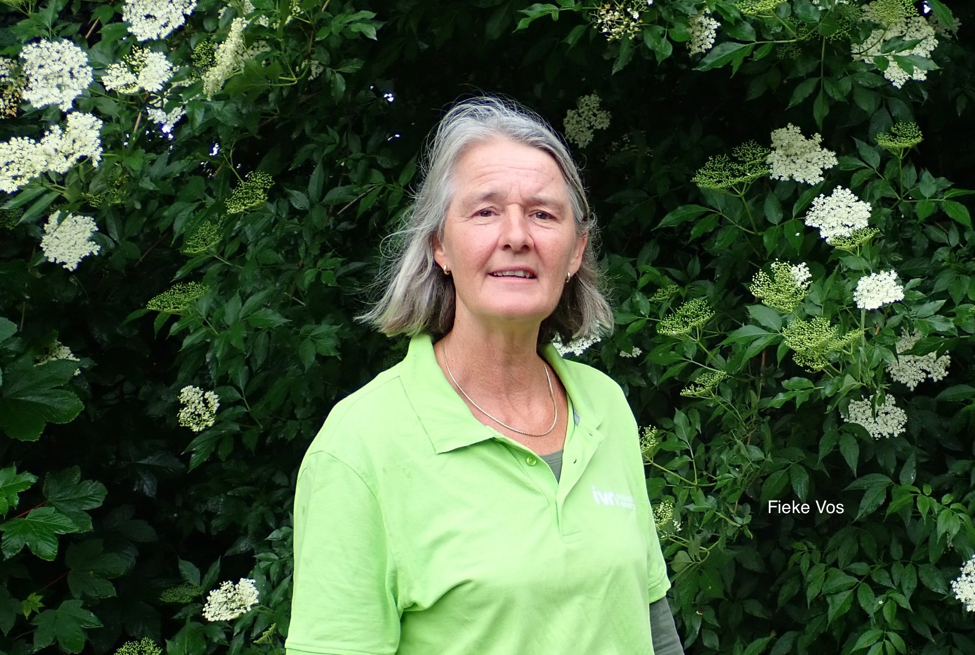 Een vrouw in een groene polo staat voor bloeiende struiken.