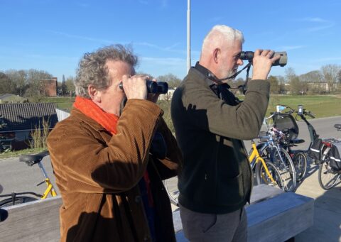 Twee mannen kijken door verrekijkers, met fietsen op de achtergrond, op een zonnige dag buiten.