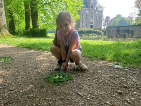 Meisje knielt op een bospad met bladeren en bloemetjes; kasteel op de achtergrond.