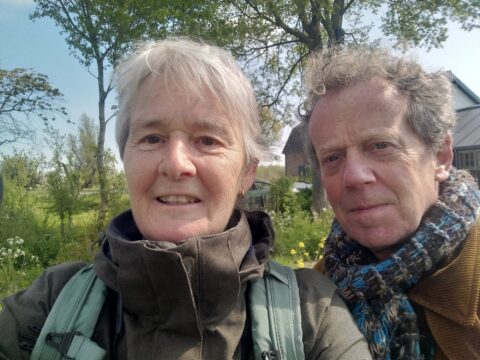 Twee mensen poseren buiten op een zonnige dag met bomen en een gebouw op de achtergrond.