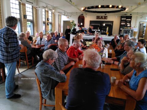 Een groep oudere mensen zit aan tafels in een café, in gesprek met elkaar.