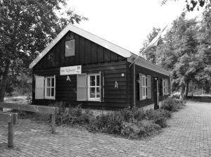 Een zwart-wit foto van een houten gebouw genaamd "Het Egboetje" met een vlag, omgeven door bomen.