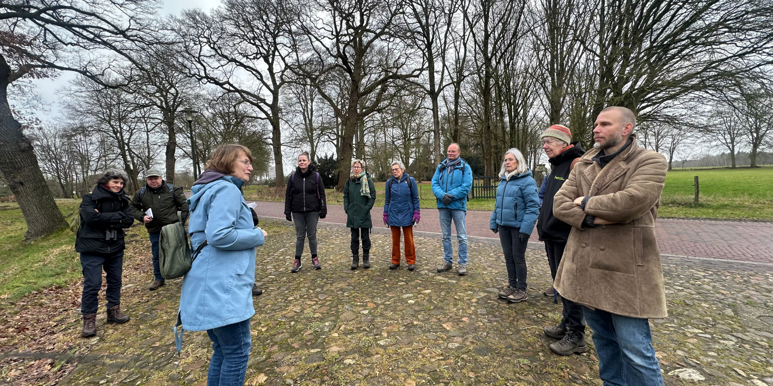 Een groep mensen staat buiten op een stenen pad omgeven door kale bomen en gras.
