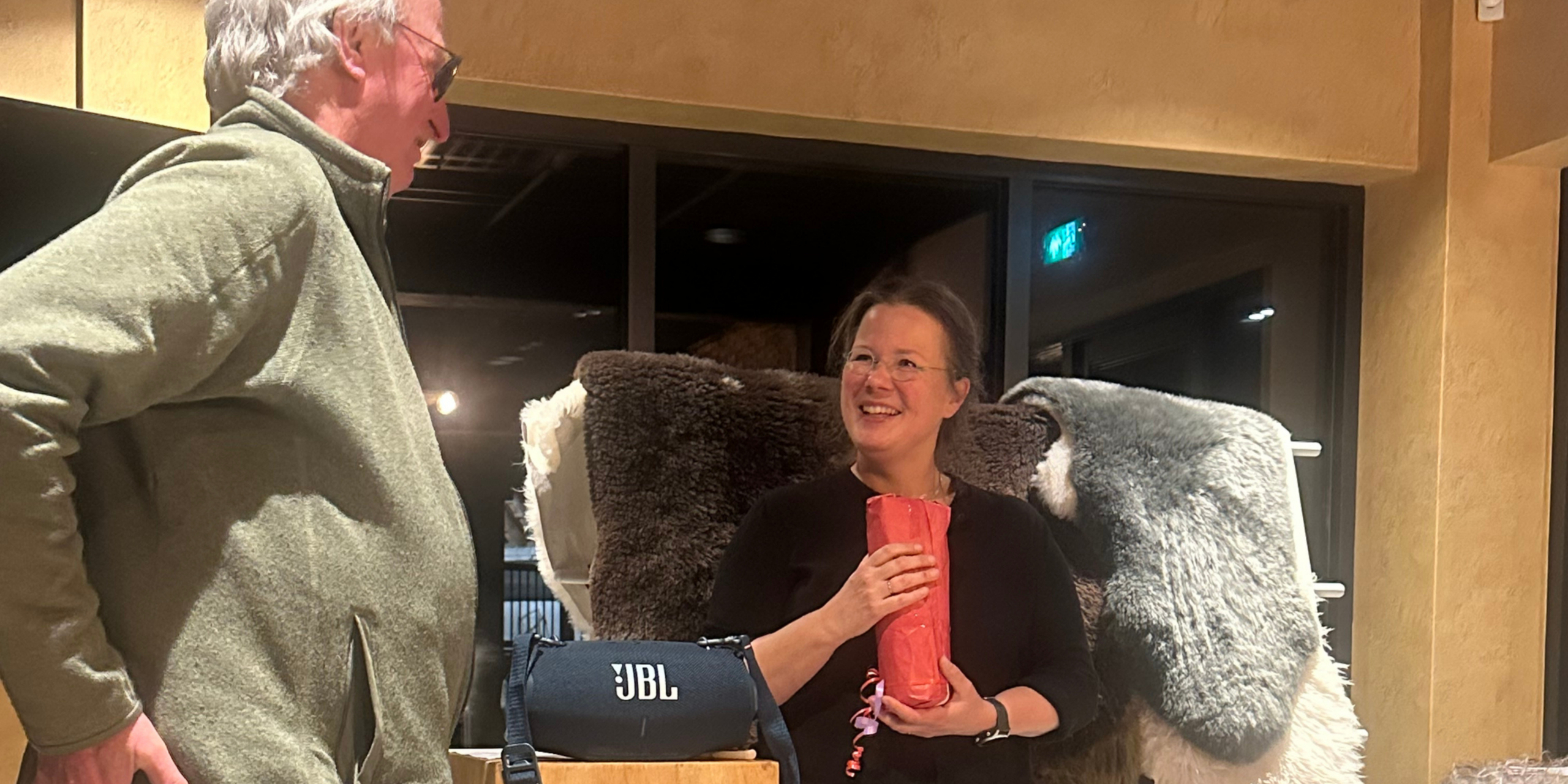 Twee mensen praten binnen; één houdt een ingepakt cadeau vast, een JBL speaker op tafel.