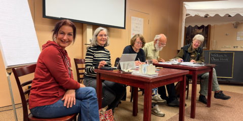 Groep mensen zit aan een tafel en schrijft, met een bord "Welkom in Oens Huus" op de achtergrond.