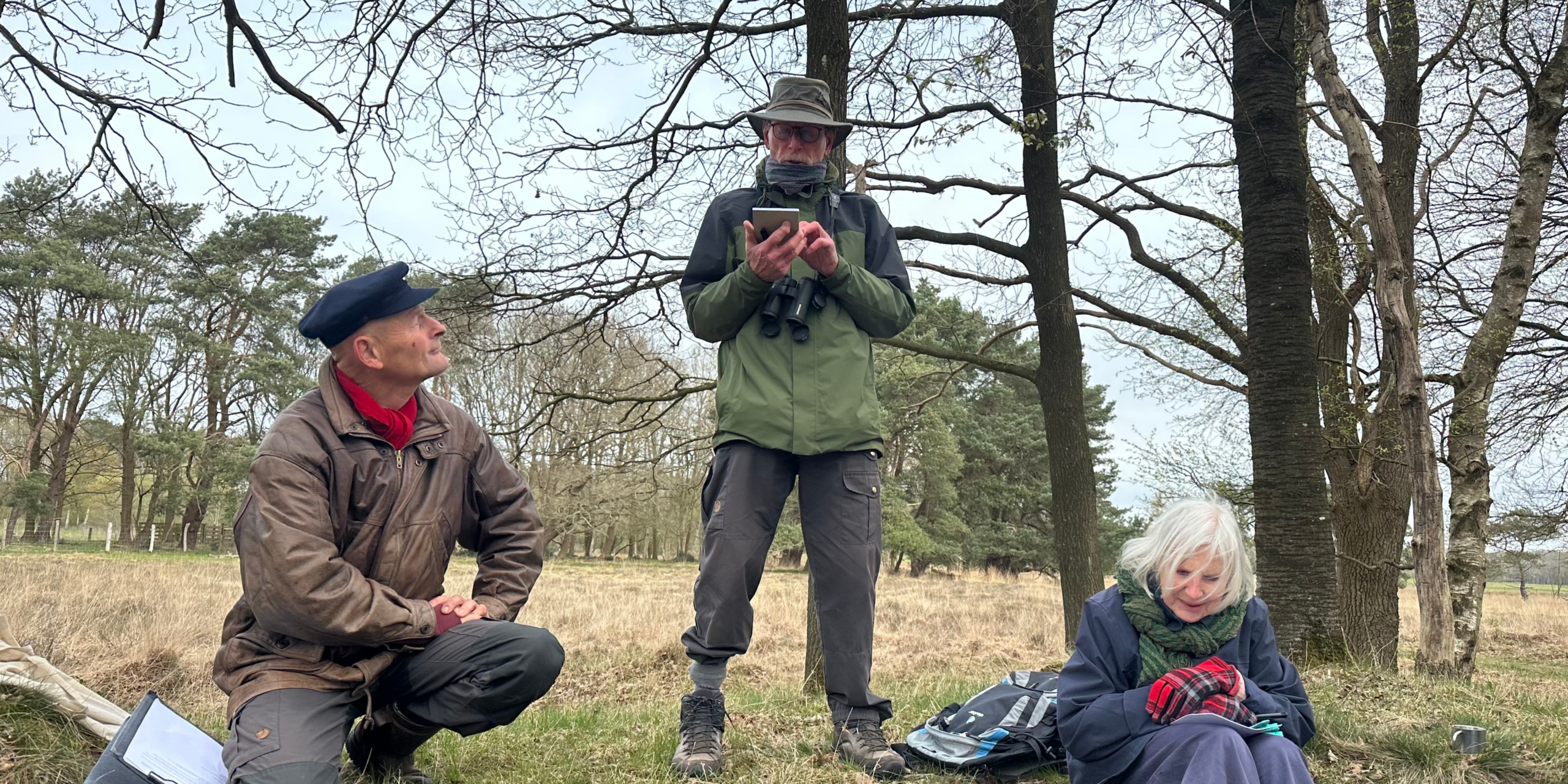 Drie mensen in een bosrijke omgeving; één hurkt, één staat met een telefoon en één zit met handschoenen.