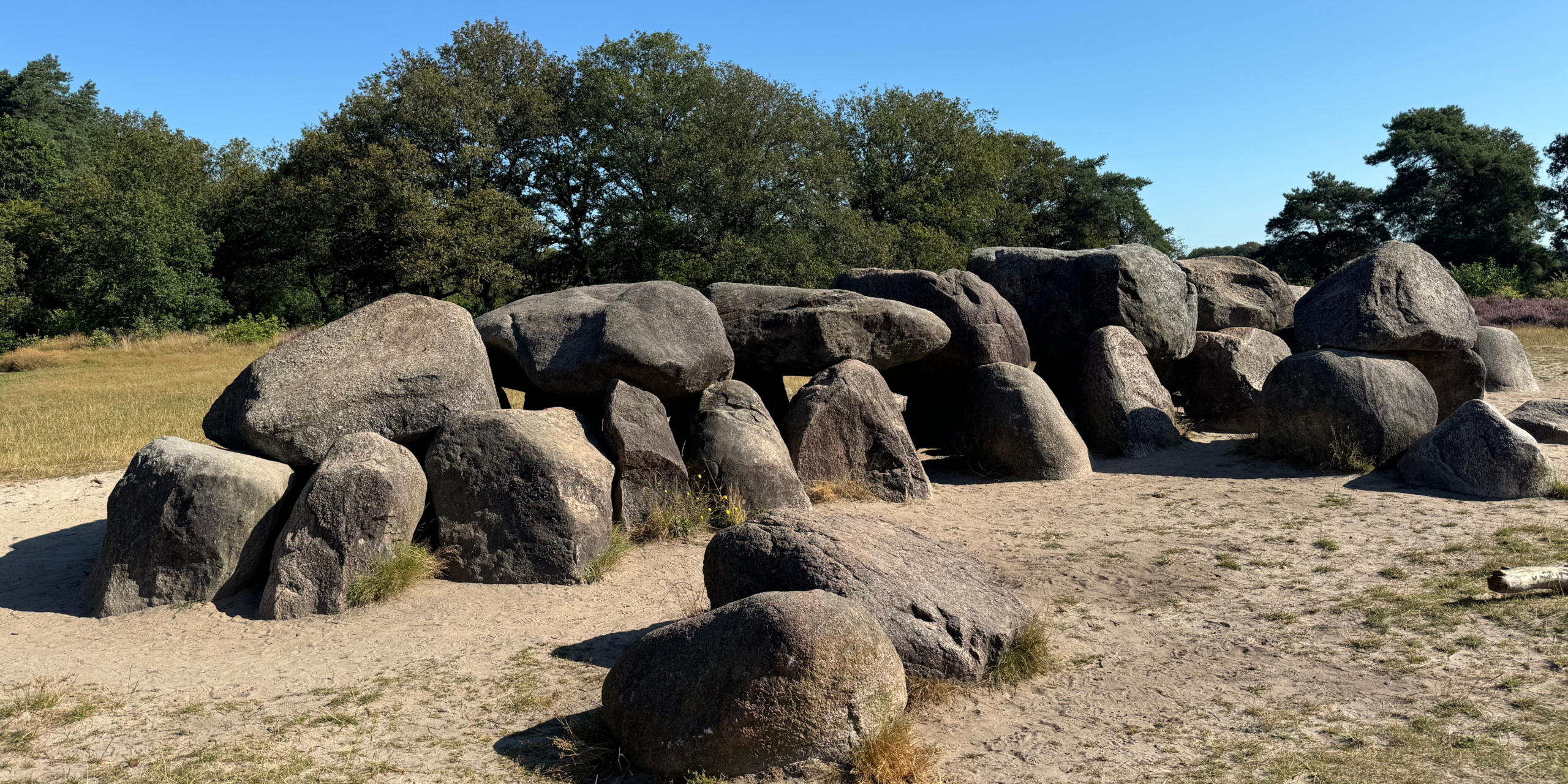 Grote zwerfstenen vormen een prehistorische hunebed in een bosrijke omgeving onder een blauwe hemel.