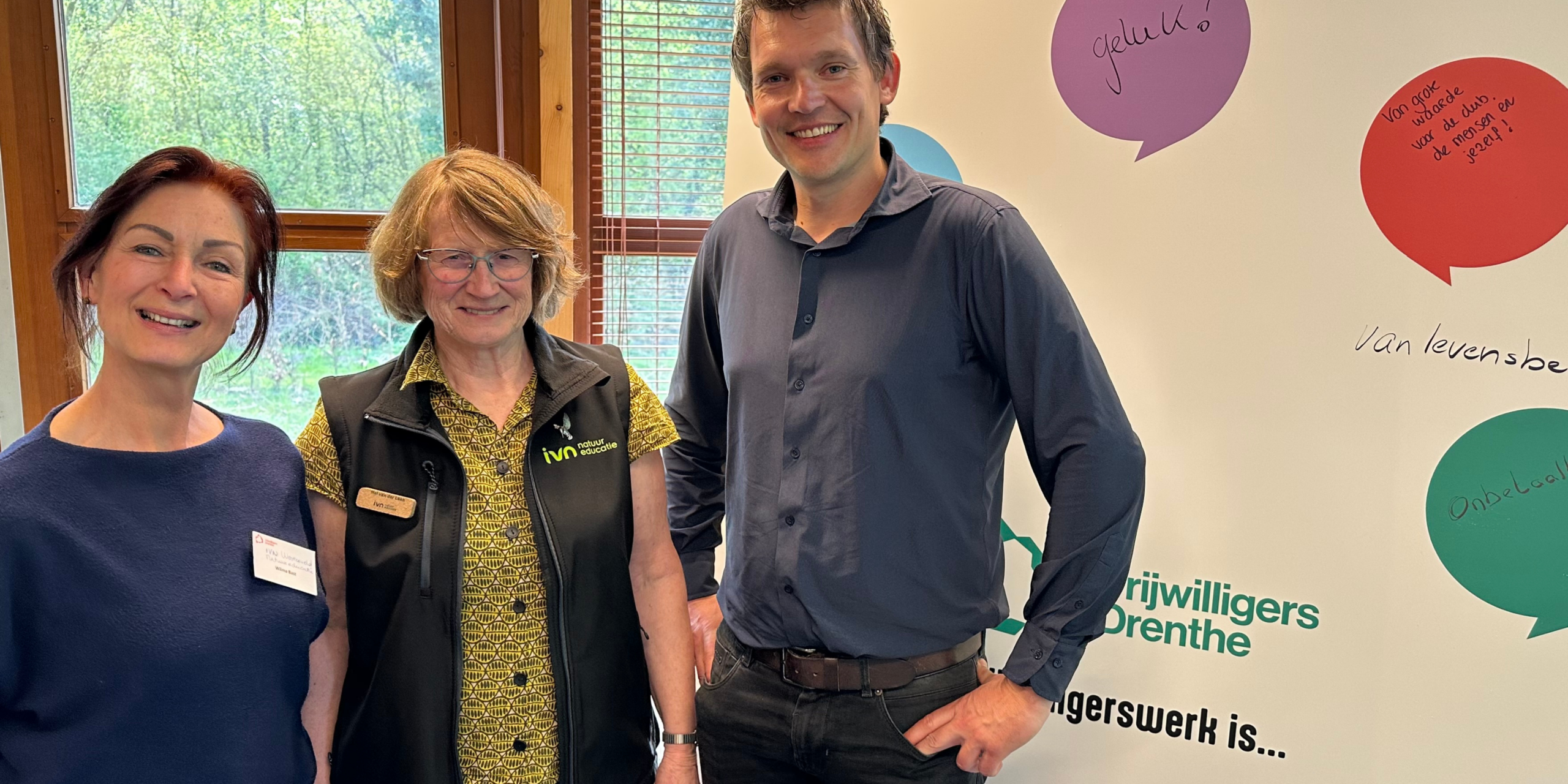 Drie mensen poseren voor een banner met tekstballonnen en "Vrijwilligers Drenthe".