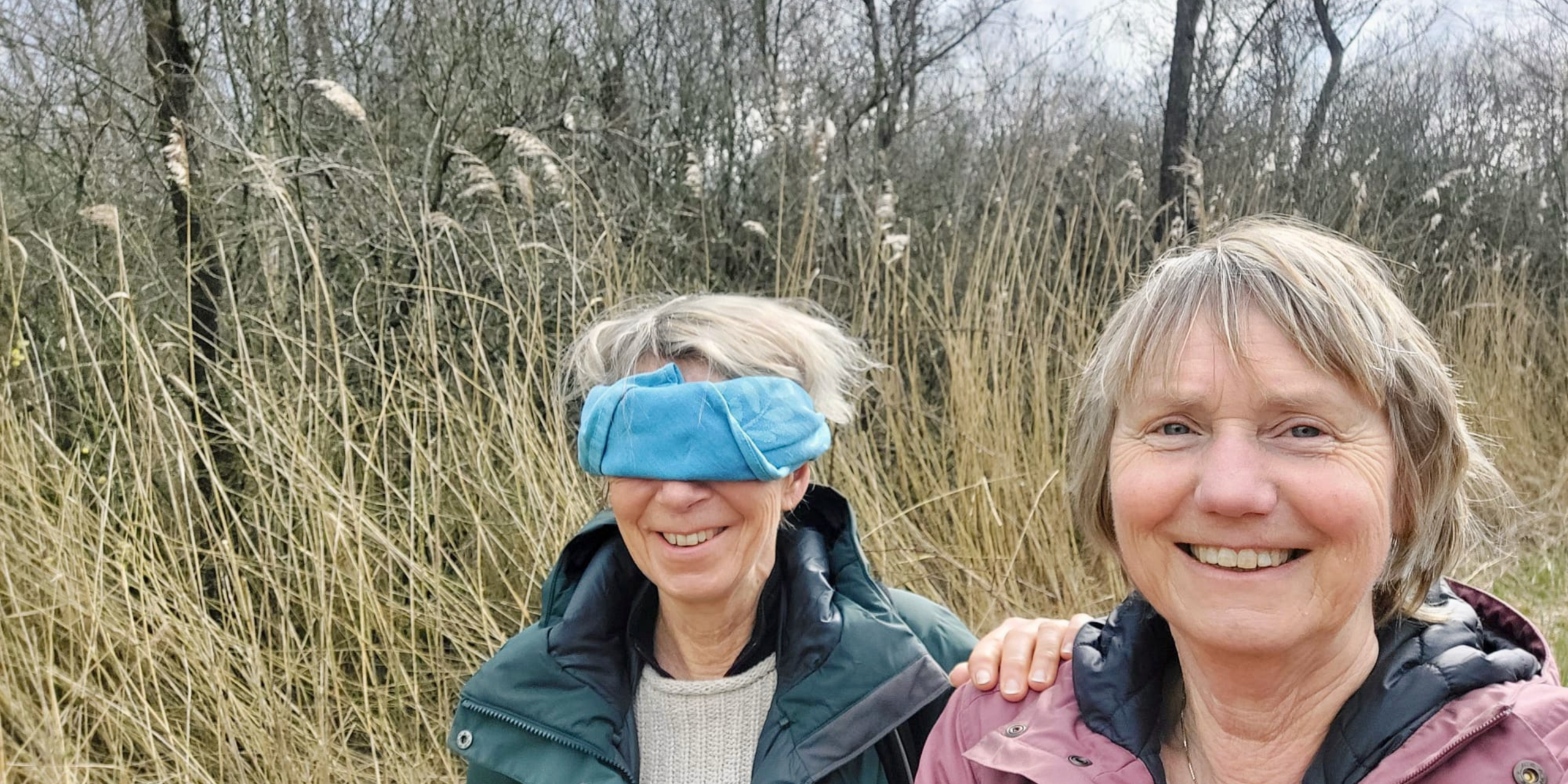 Twee lachende vrouwen in een grasland, één geblinddoekt met blauw doek.