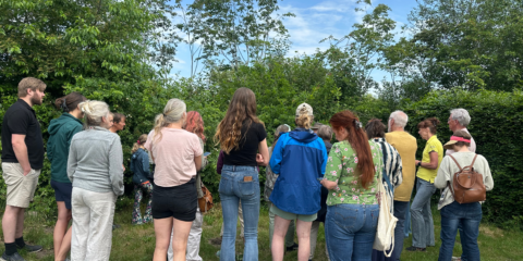 Groep mensen in casual kleding luistert naar een spreker bij groen gebladerte.