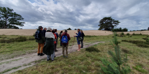 Groep wandelaars luistert naar gids in graslandschap met zandheuvels en wolkenlucht.