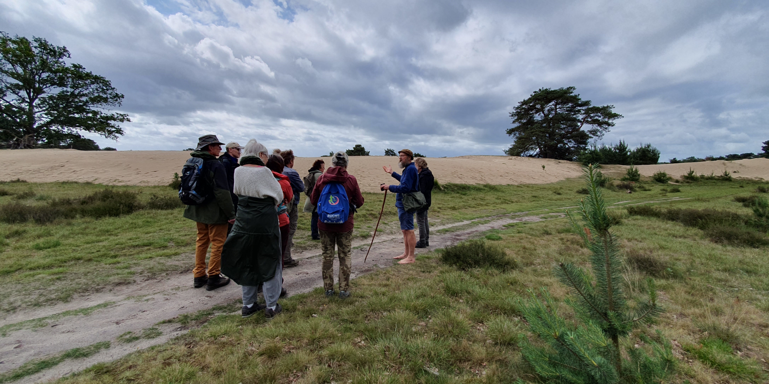 Groep wandelaars luistert naar gids in graslandschap met zandheuvels en wolkenlucht.