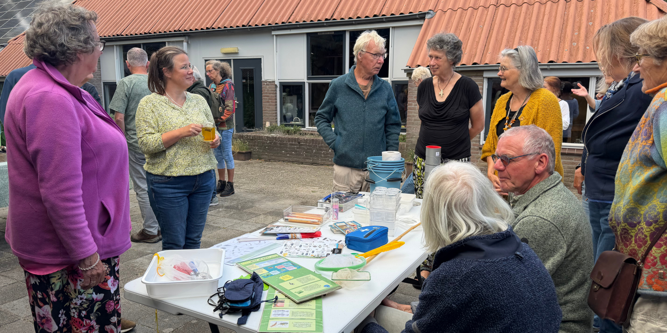 Groep mensen rond een tafel buiten, met informatiemateriaal en drinken. Gezellig gesprek.