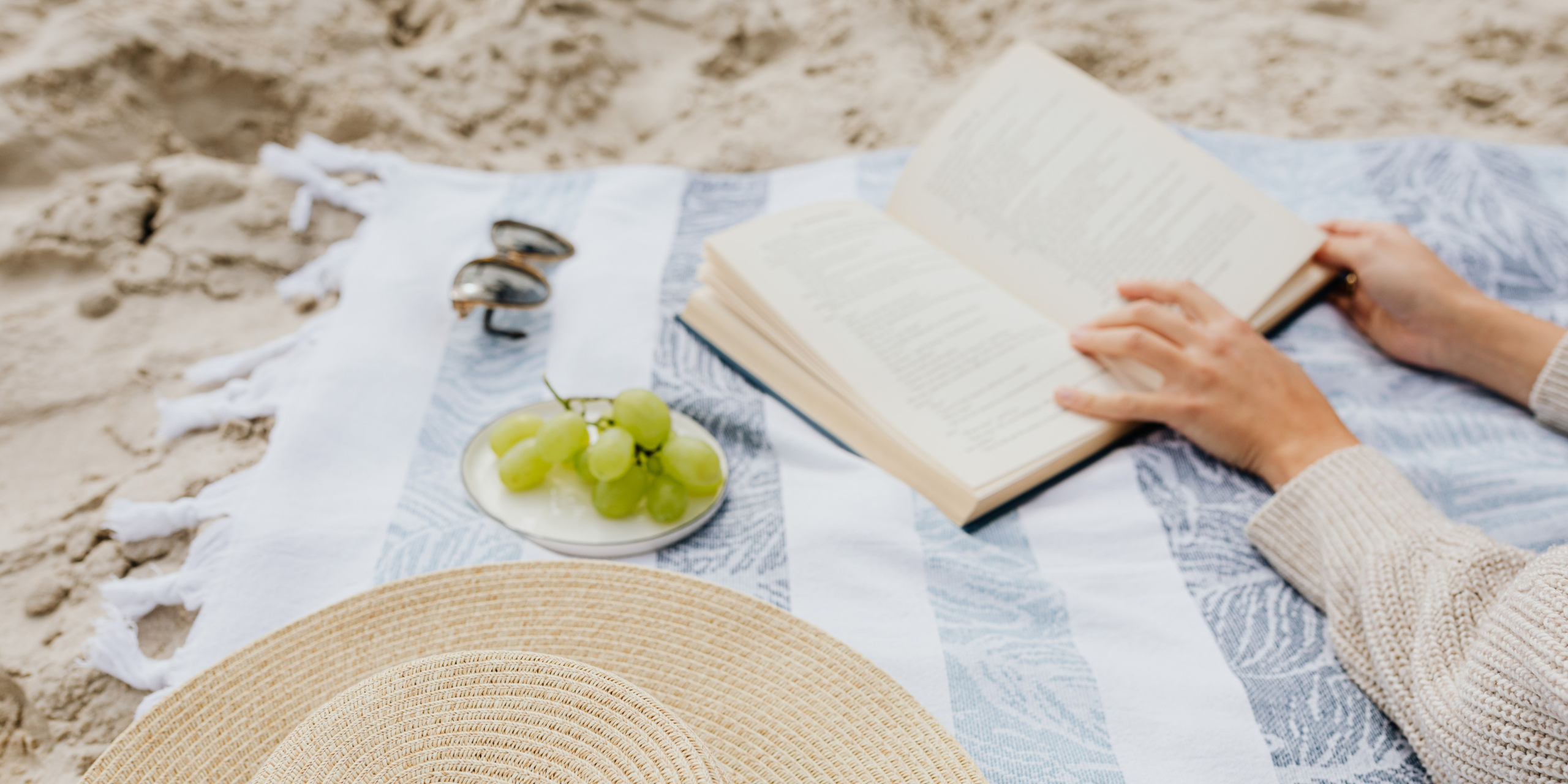 Persoon leest een boek op een strandlaken met een strohoed, druiven en zonnebril.