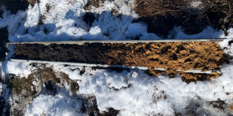 Grondkernmonster in metalen houder op sneeuw, met donkere en lichtere bodemlagen zichtbaar.