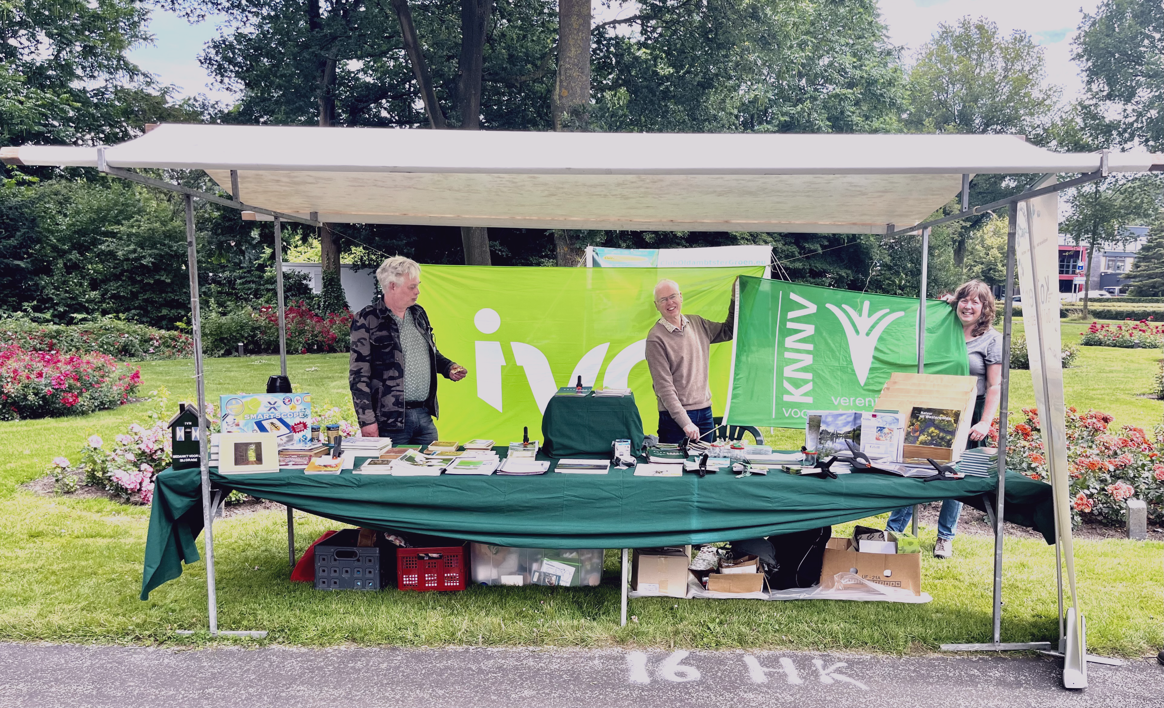 Drie mensen bij een informatiestand met groene IVN- en KNNV-vlaggen in een park.